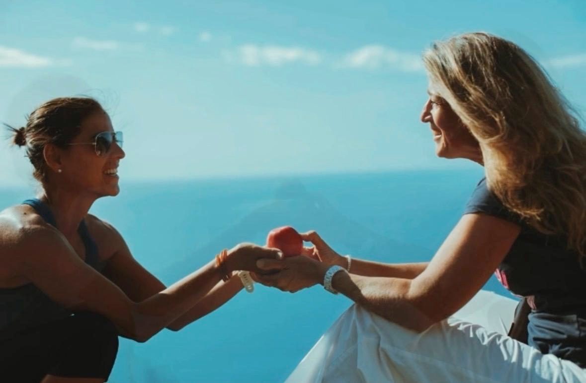 Zwei Frauen auf einer Terrasse über dem Meer reichen
einander lächelnd eine rote Frucht — das
Fastenbrechen