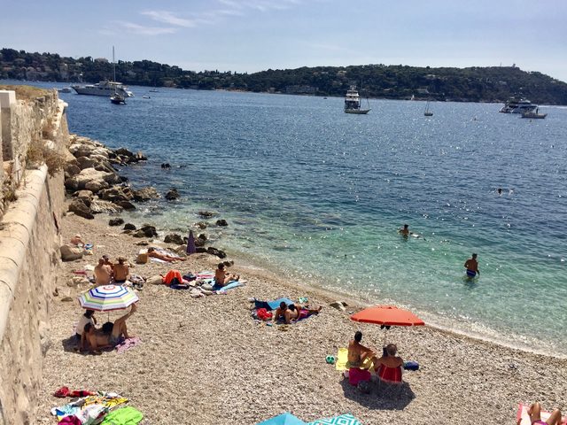 Kiesstrand in einer Bucht mit Sonnenschirmen, Badegästen und ankernden Booten